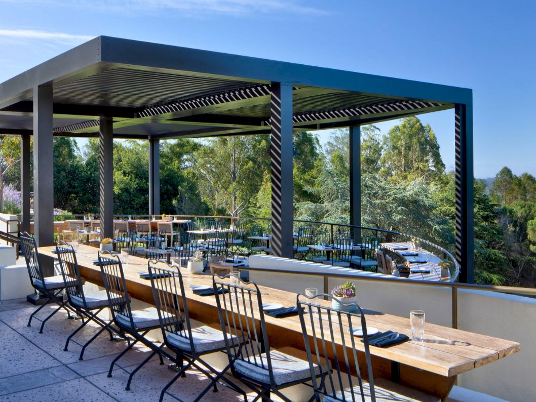 A stylish outdoor terrace with wooden tables and iron chairs, shaded by a modern pergola, overlooking trees and blue sky.