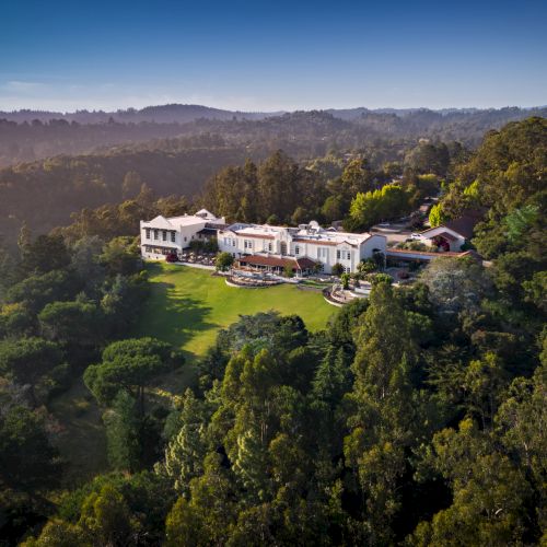 Luxurious mansion complex nestled in dense forest, white buildings with manicured lawn surrounded by tall trees and distant hills.