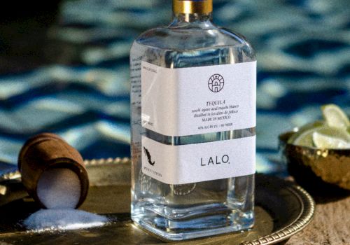 A bottle of LALO tequila sits on a decorative tray with a chocolate scoop, lime bowl, and a blurred pool background&mdash;simple luxury.