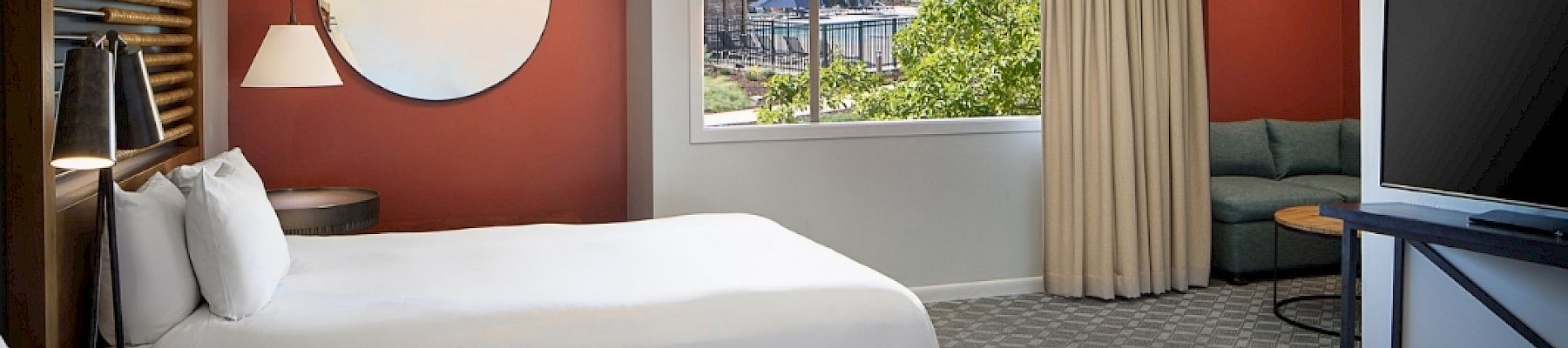 A hotel room with two beds, a large TV, a round mirror, a window with a view, and a small sitting area with a couch and table.