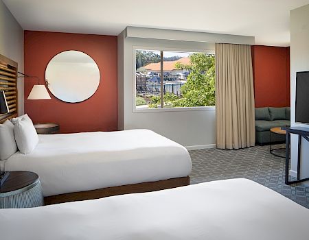 A hotel room with two beds, a large TV, a round mirror, a window with a view, and a small sitting area with a couch and table.