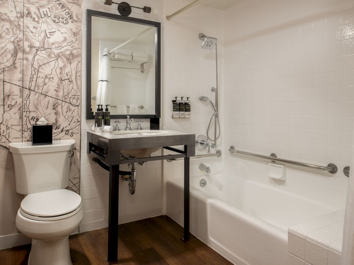 A modern bathroom with a white toilet, a black-framed mirror above a dark vanity, a white tub with a handheld shower, grab bar, and a map-patterned accent wall.