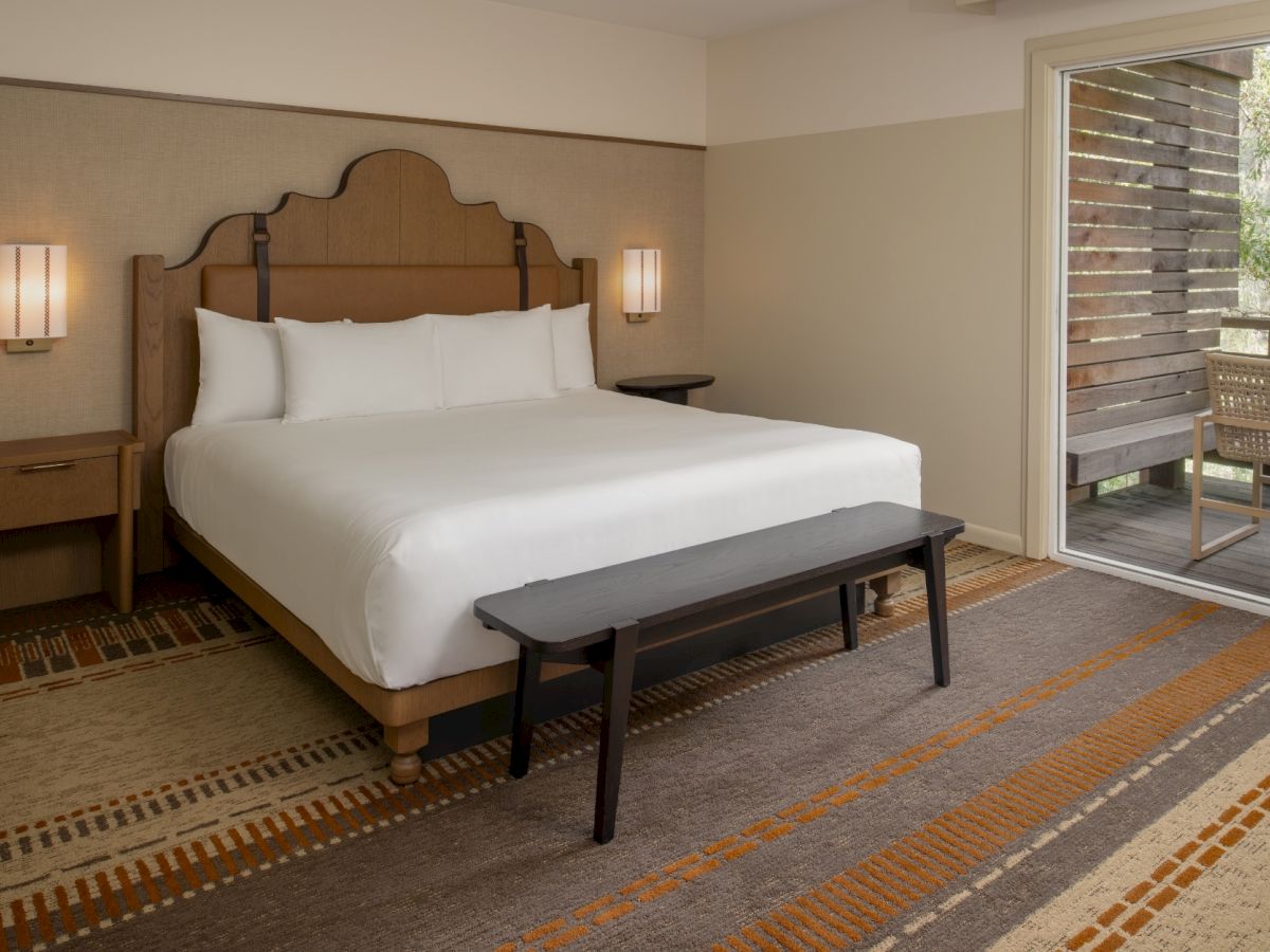 A hotel room with a king bed, nightstands, bench at foot, warm lighting, and a sliding door to a balcony. End.