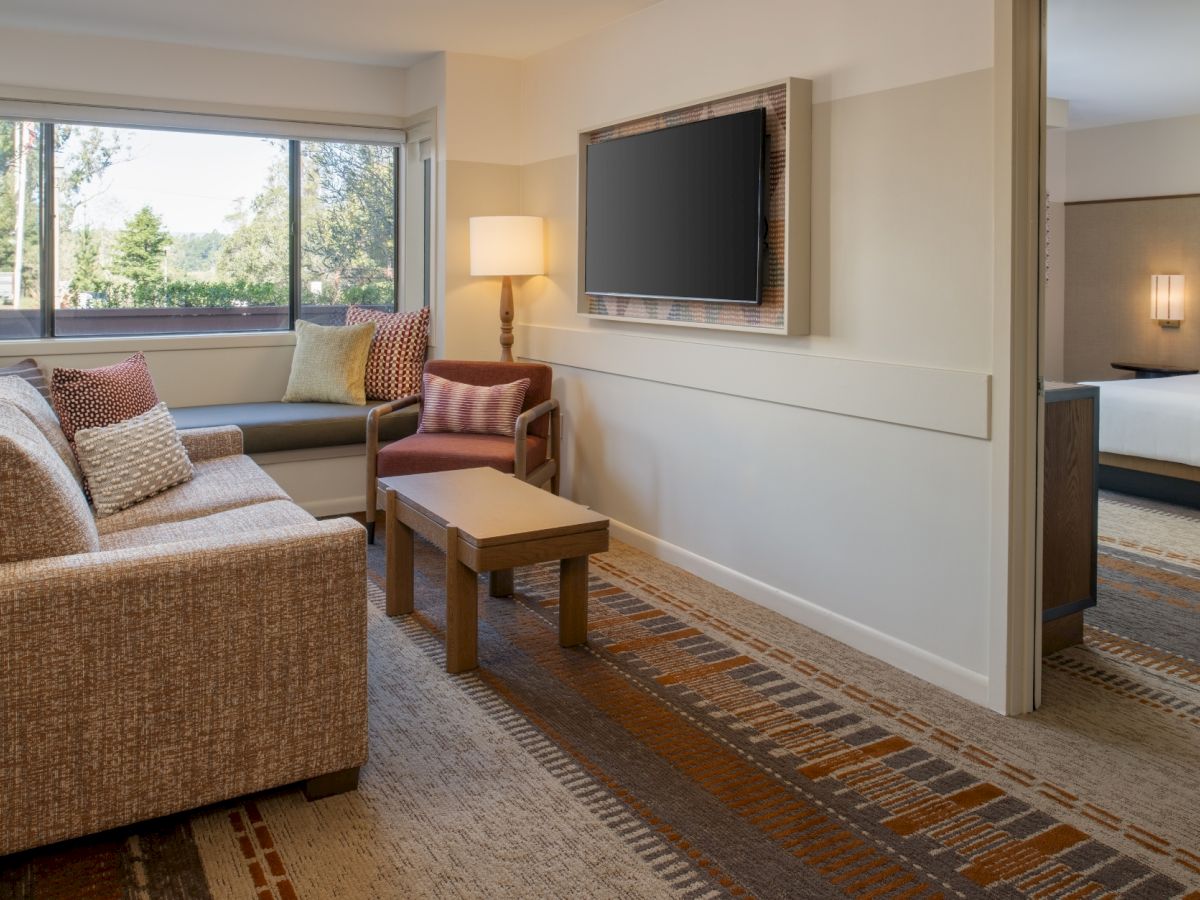 A cozy hotel suite living area with a beige sofa, brown chairs, coffee table, wall-mounted TV, large window, and soft lighting; door to a bedroom in the background.