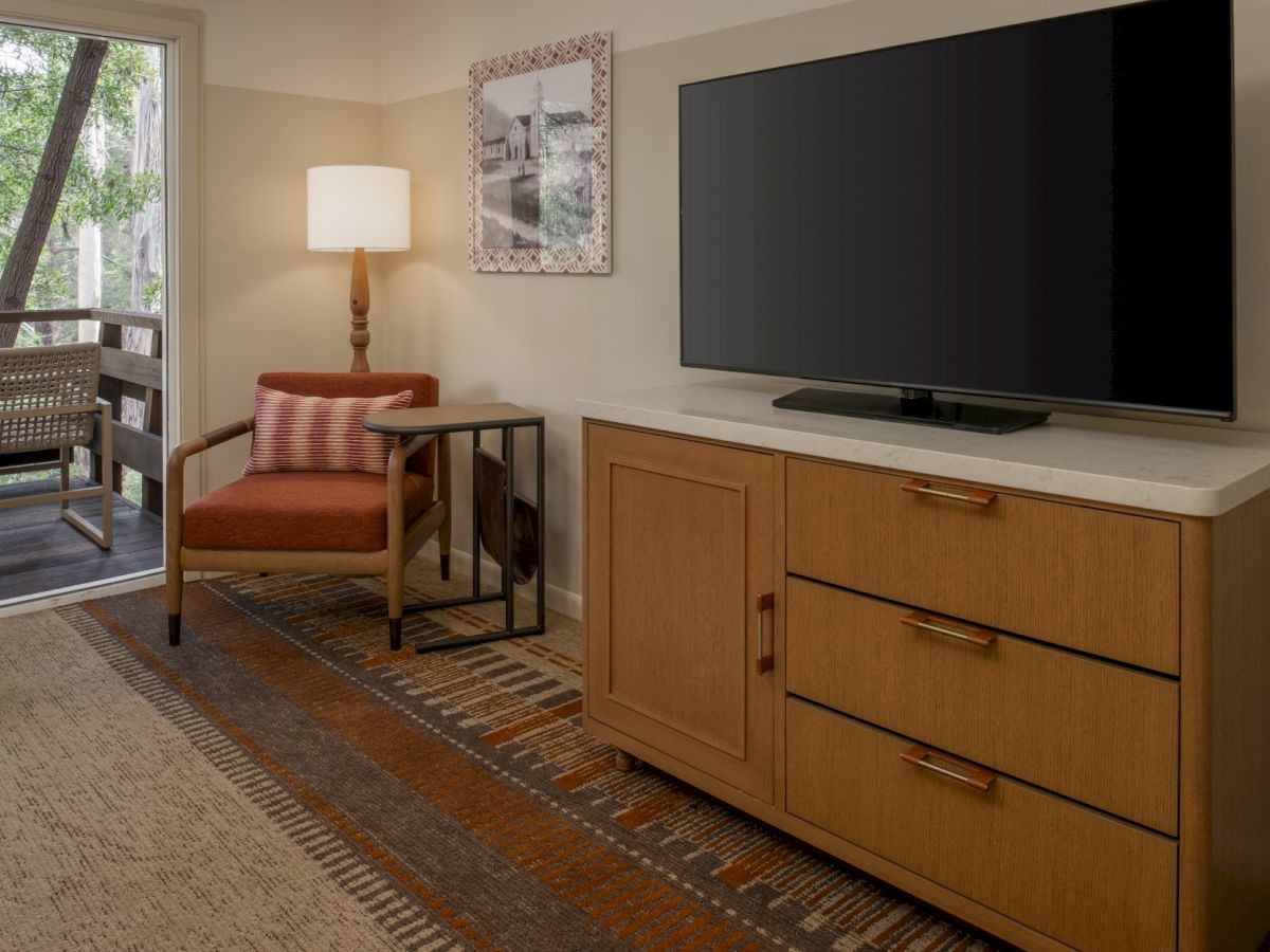 A cozy hotel room with a flat-screen TV on a wooden dresser, a seating chair, lamp, and a doorway to a balcony.