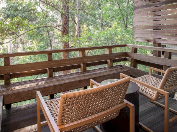 A wooden balcony with woven chairs around a small table, built-in railing, and trees visible beyond the railing, shaded by a wooden wall.