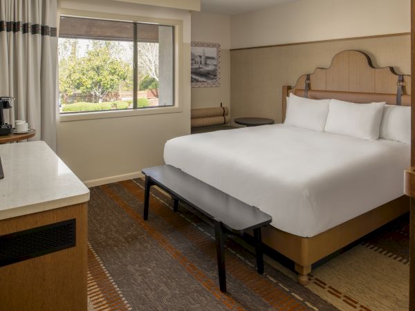 A clean hotel room with a large bed, white linens, a bench at the foot, a desk with a TV, a window with curtains, and a chair by the wall.