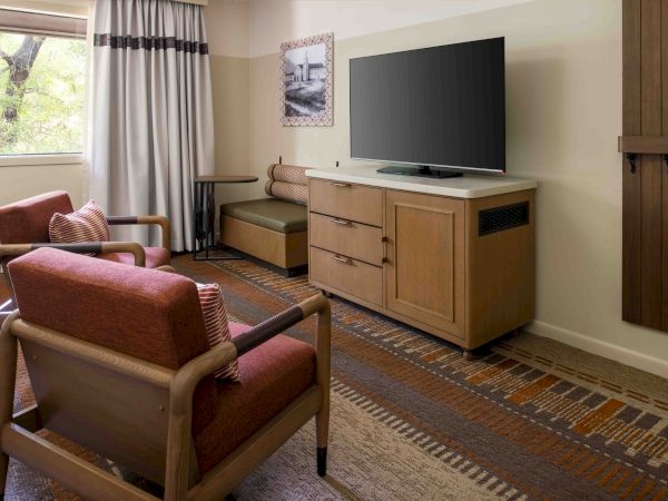 A cozy hotel living area with two red-armchair chairs, a wooden TV stand, a mounted flat-screen TV, and a window with curtains.