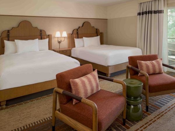 Two beds in a hotel room with two red armchairs and a small table between them, plus a balcony outside.
