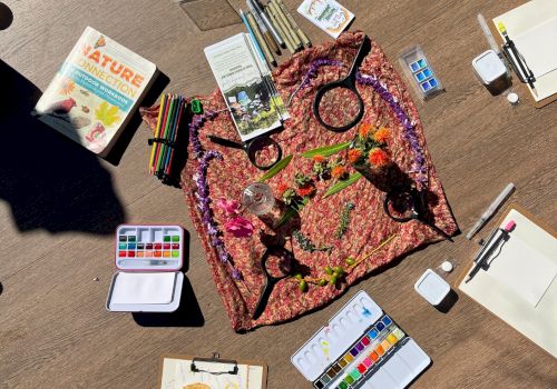 Colorful art supplies arranged around a knotted, tie-dye-like fabric on a wooden surface, with paints, brushes, and sketchbooks scattered about.