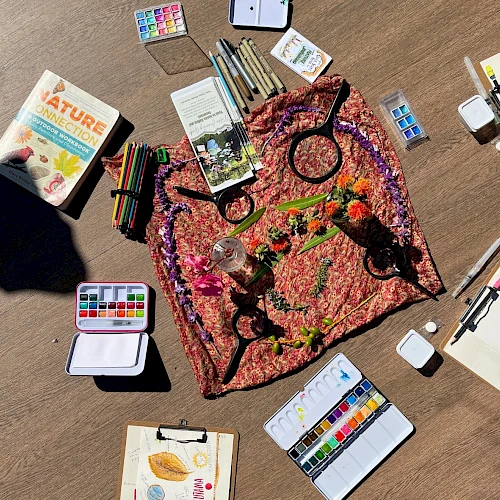 Colorful art supplies arranged around a knotted, tie-dye-like fabric on a wooden surface, with paints, brushes, and sketchbooks scattered about.