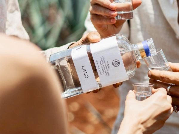 People pouring clear liquor into shot glasses from a bottle labeled &ldquo;ALQ&rdquo; during a group drink. 140 chars ending with a period.