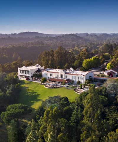 An aerial shot of a large, luxurious mansion nestled in a forested area with expansive lawn and greenery surrounding the property, ending the sentence.