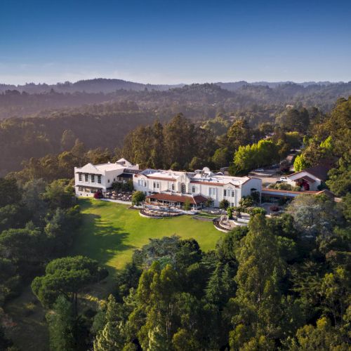 An aerial shot of a large, luxurious mansion nestled in a forested area with expansive lawn and greenery surrounding the property, ending the sentence.