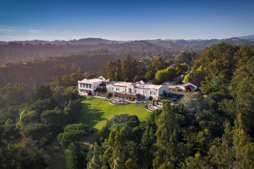 An aerial shot of a large, luxurious mansion nestled in a forested area with expansive lawn and greenery surrounding the property, ending the sentence.