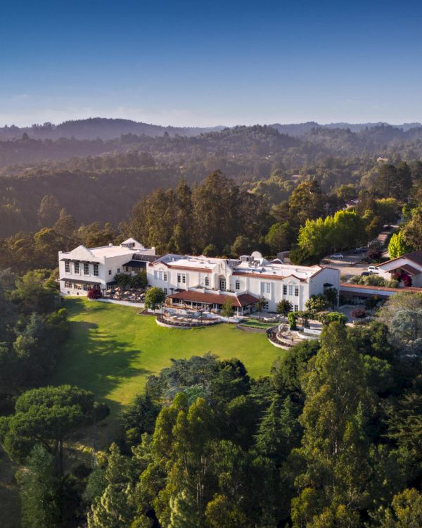 An aerial shot of a large, luxurious mansion nestled in a forested area with expansive lawn and greenery surrounding the property, ending the sentence.