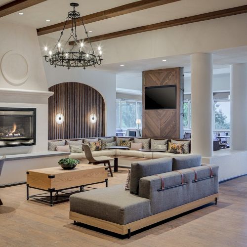 A spacious, modern living area with a large fireplace, seating arrangements, wooden flooring, a chandelier, and a wall-mounted TV.