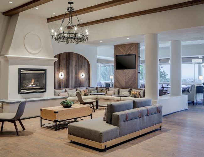 A spacious, modern living area with a large fireplace, seating arrangements, wooden flooring, a chandelier, and a wall-mounted TV.