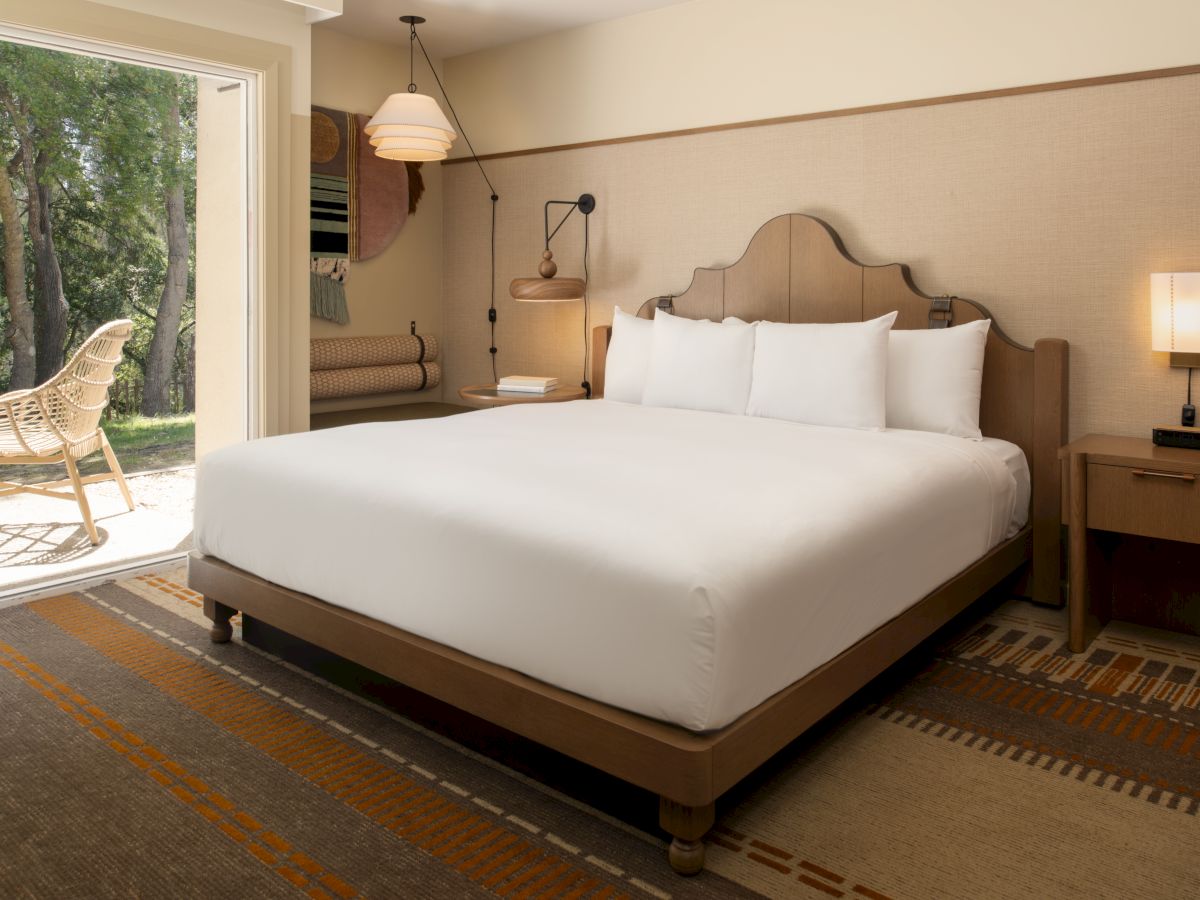 A cozy hotel room with a king bed, nightstands, lamps, and a sliding door opening to a garden patio. The scene feels bright and welcoming.