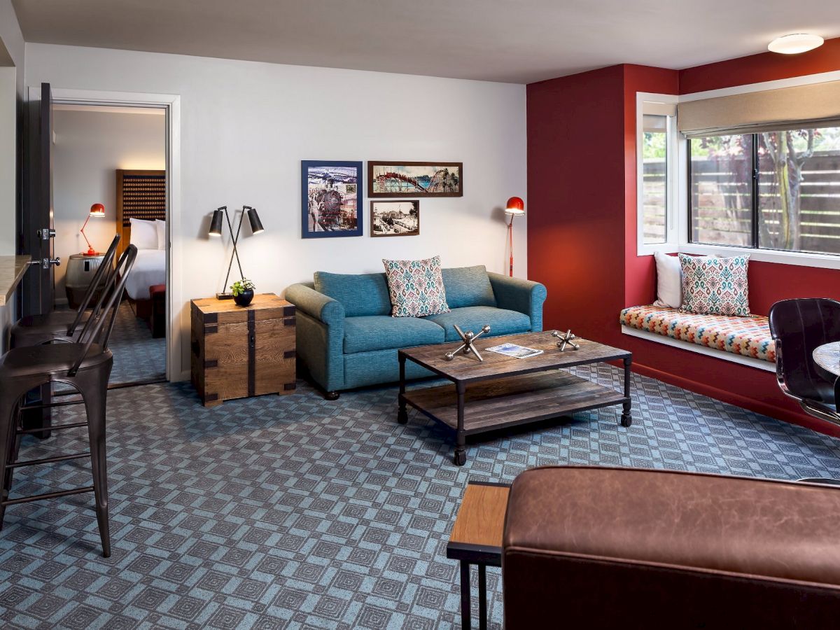 The image shows a living room with blue furniture, a window seat, a coffee table, framed pictures on the wall, and adjoining bedroom viewed from open door.