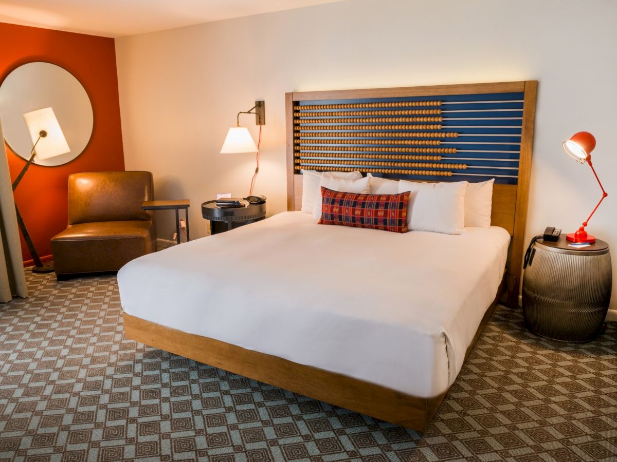 A modern hotel room with a large bed, decorative headboard, armchair, two lamps, a circular mirror, and a patterned carpet.