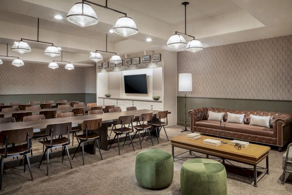 A modern conference room with rows of desks and chairs, a TV on the wall, a leather sofa, a coffee table, and green ottomans for seating.