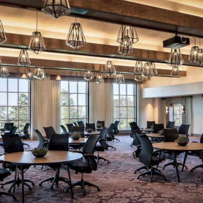 The image shows a spacious, well-lit conference room with round tables, surrounded by office chairs, modern light fixtures, and large windows.