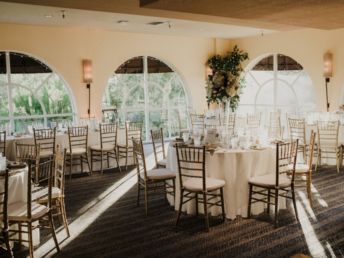 A banquet hall with round tables, elegantly set for an event, featuring tall windows, draped chairs, and floral centerpieces, ready for guests.