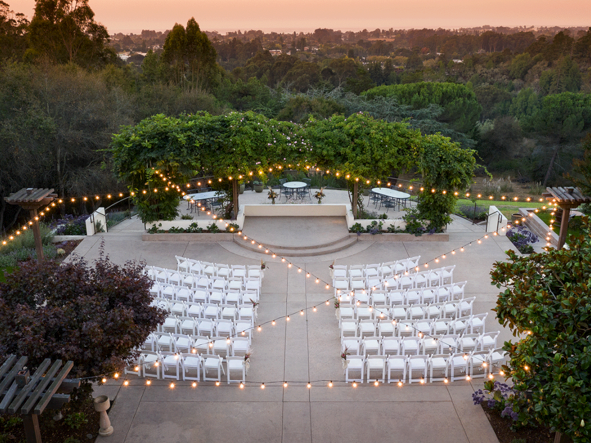 An outdoor wedding setup features white chairs arranged in rows, string lights overhead, and a greenery-covered pergola with a scenic sunset backdrop.