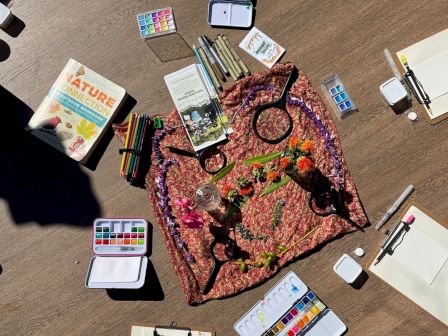 A watercolor palette, brushes, and art supplies arranged around a round rug on a wooden floor, with painted plants and jars.