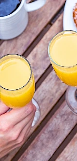 Two hands holding glasses of orange juice over a wooden table set with various breakfast foods, including pastries and coffee.