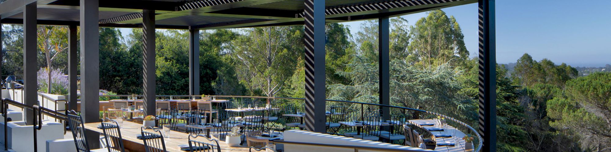 An outdoor restaurant patio with long wooden tables and black chairs, under a modern pergola structure, surrounded by greenery and a clear sky.