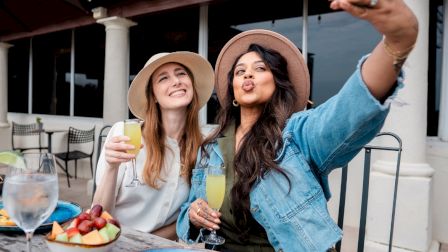 Two friends taking a selfie on a sunny patio, toasting drinks with colorful plates of food in front of them, smiling and posing.