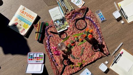 A colorful arts-and-crafts scene with paints, brushes, notebooks, and a patterned fabric bag spread on a wooden table, colorful supplies scattered around.
