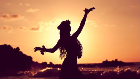 A silhouette of a person dancing on the beach at sunset, arms raised, with waves and rocks in the background.
