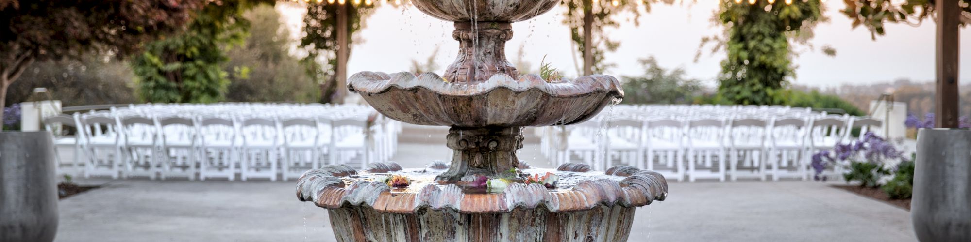 A decorative fountain is in the foreground, in front of outdoor seating and string lights under a canopy of greenery at dusk.