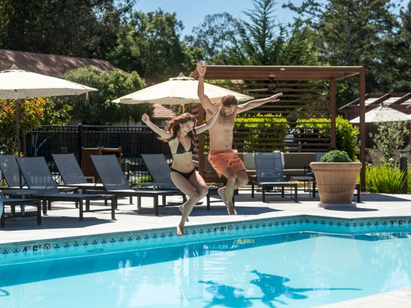 Two people joyfully jump into a pool from the edge, arms raised, with lounge chairs and umbrellas around a sunny resort pool area.