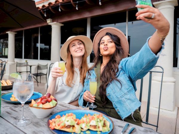 Two friends in sun hats selfie at an outdoor cafe, smiling with colorful drinks and plates of food on the table.