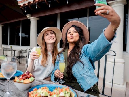 Two friends selfie-ing while enjoying drinks and food on a sunny patio, smiling and posing for a cheerful shot.