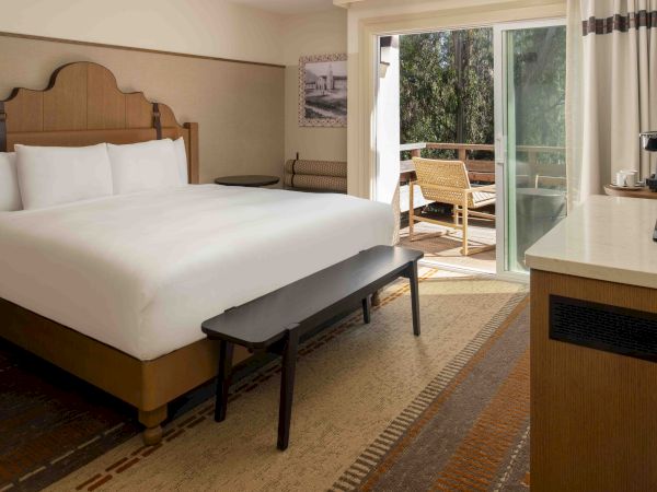 A hotel room with a large bed, bench at the foot, desk, chair, and sliding glass door opening to a balcony with another chair and greenery outside.