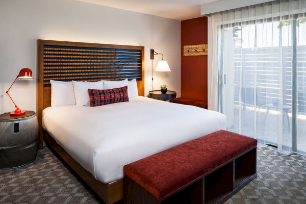 A modern hotel room with a king-size bed, red bench, bedside lamp, and sliding glass door leading to a patio.