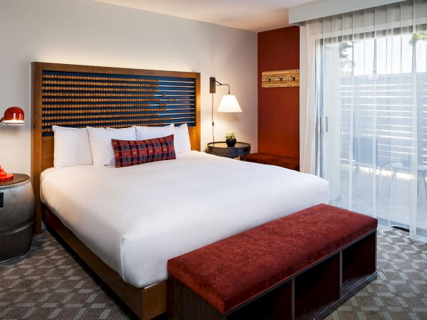 A modern hotel room with a king-size bed, red bench, bedside lamp, and sliding glass door leading to a patio.