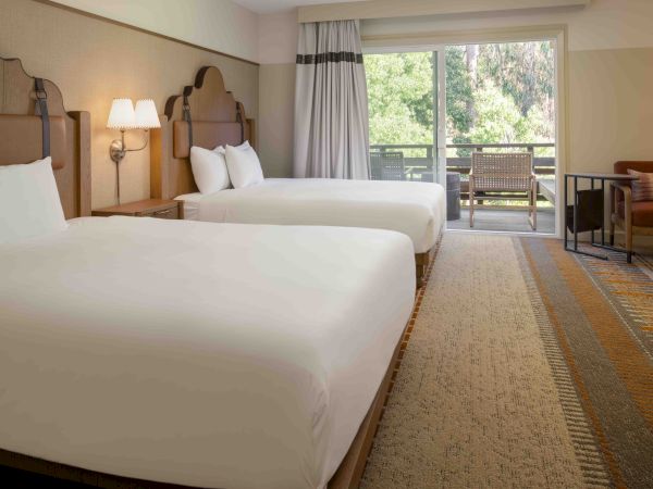 Two white-blanketed hotel beds, a bedside lamp, and a balcony with seating overlooking greenery. The room has patterned carpet and light walls.