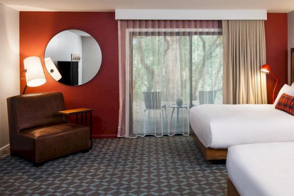 A hotel room with two beds, a brown armchair, a round mirror, and a sliding glass door leading to a balcony with outdoor seating.