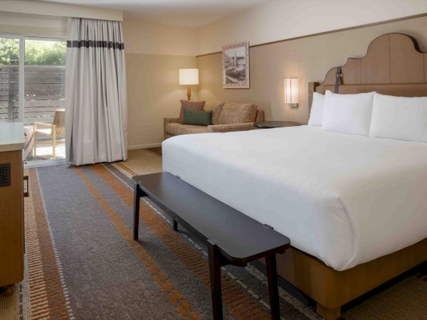 A hotel room with a large bed, neutral tones, a seating area, a desk, a bench at the foot of the bed, and a sliding door to a balcony.