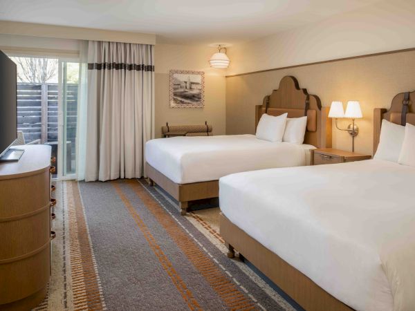 A hotel room with two queen beds, nightstands, lamps, a dresser with a TV, a sliding door to a balcony, and warm decor.