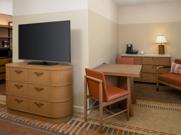 A cozy hotel suite with a large flat-screen TV on a wooden dresser, a wooden table and chairs, a sofa, and soft ambient lighting by a lamp.