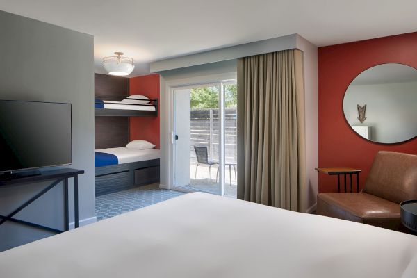 This image shows a modern hotel room with a bed, bunk beds, a TV, a sliding glass door to a patio, a round mirror on a red wall, and a chair.