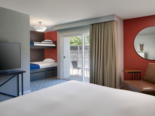 This image shows a modern hotel room with a bed, bunk beds, a TV, a sliding glass door to a patio, a round mirror on a red wall, and a chair.