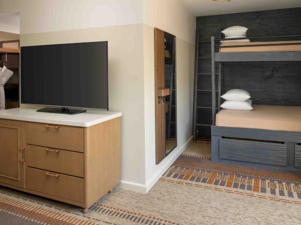 A hotel room with a dresser and a large flat-screen TV, a mirror, and a bunk-style bed with dark wood and light bedding, plus a closet area.